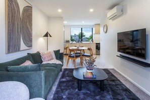 A modern living room with a green sofa, a coffee table with a book and a plant, a dining area with wooden chairs and a table, and a TV mounted on the wall. - Luxury CBD Retreat | 2 King Beds + Ensuites (Christchurch)