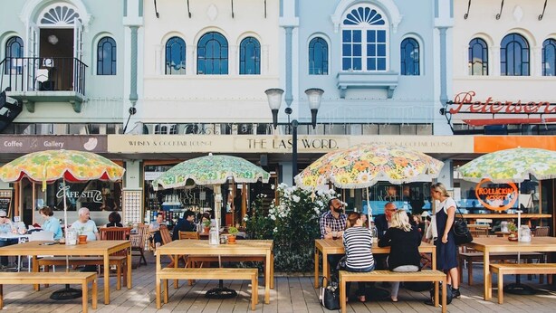 Een terras met houten tafels en banken, omringd door gebouwen met verschillende winkels en een zichtbaar bord van café 'The Last Word'.