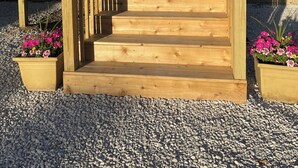 Una escalera de madera que lleva a una puerta, con una antena parabólica arriba y macetas con flores en la base.