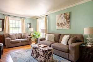 Ein Wohnzimmer mit zwei Sofas, einem gemusterten Hocker, einem Couchtisch, einer Lampe, einer Pflanze und einem Gemälde von einem Pferd an der Wand.