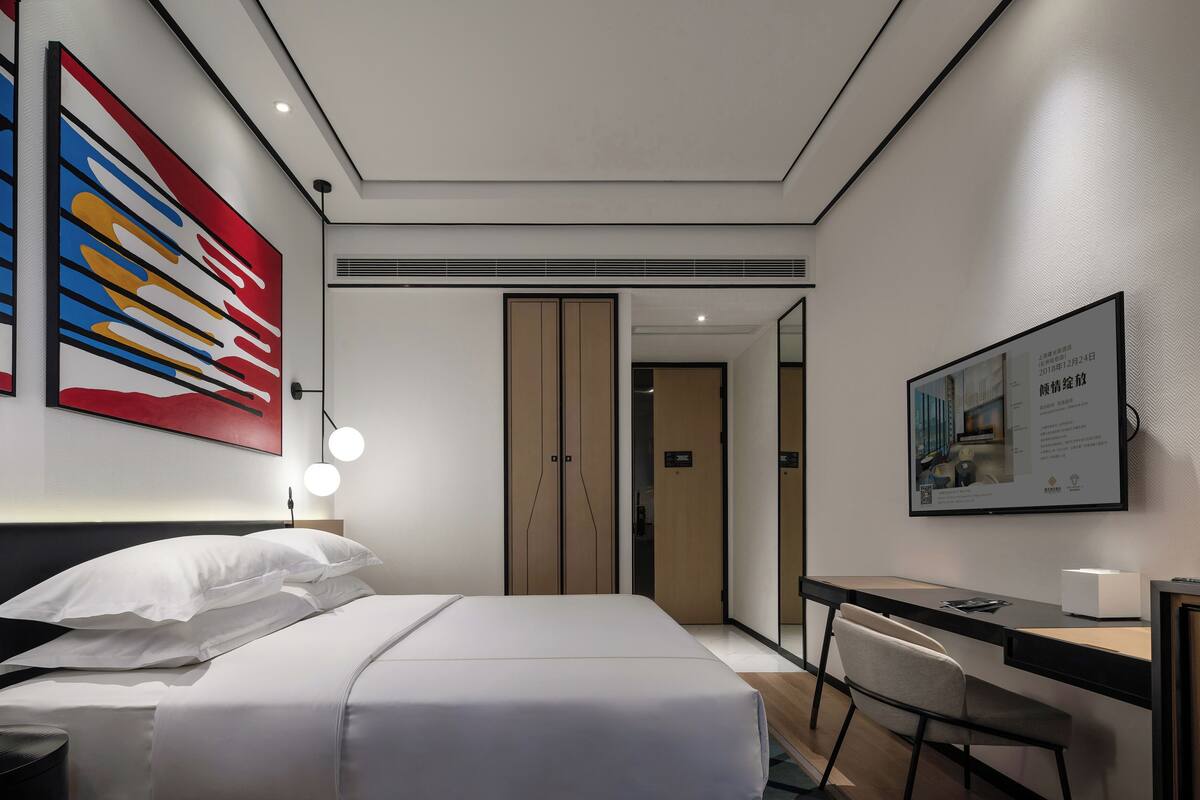 A modern hotel room with a neatly made bed, a desk with a chair, a large artwork on the wall, and a framed picture on the desk.