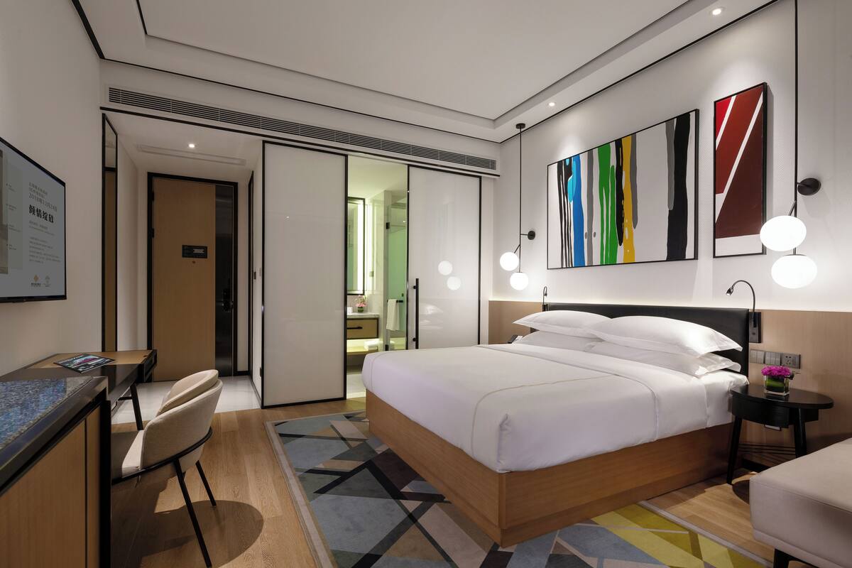 A modern hotel room with a bed, desk, chair, and artwork on the wall.