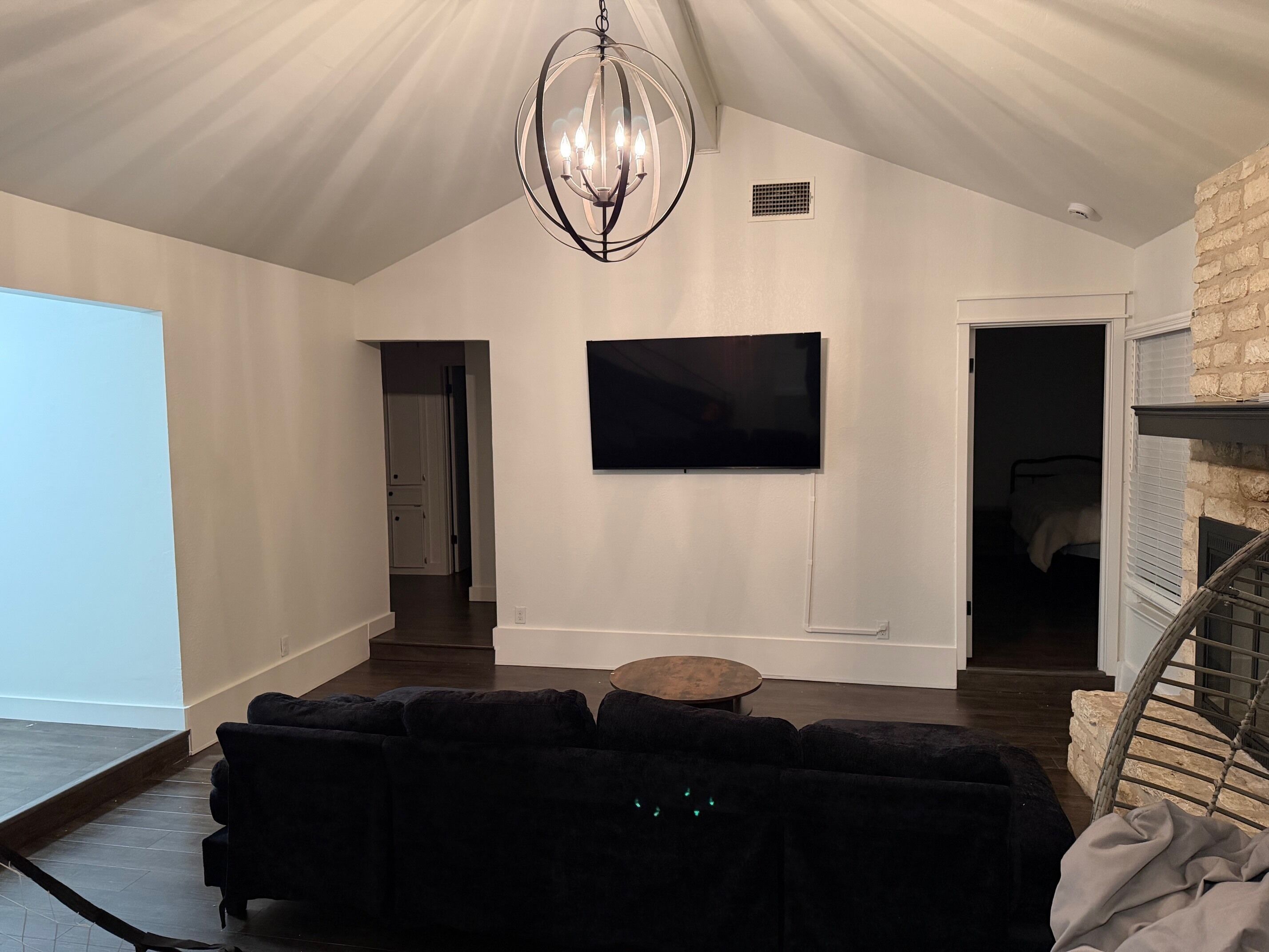 Un salon moderne avec un canapé d’angle noir, une table basse ronde, un grand téléviseur à écran plat fixé au mur et un lustre original.