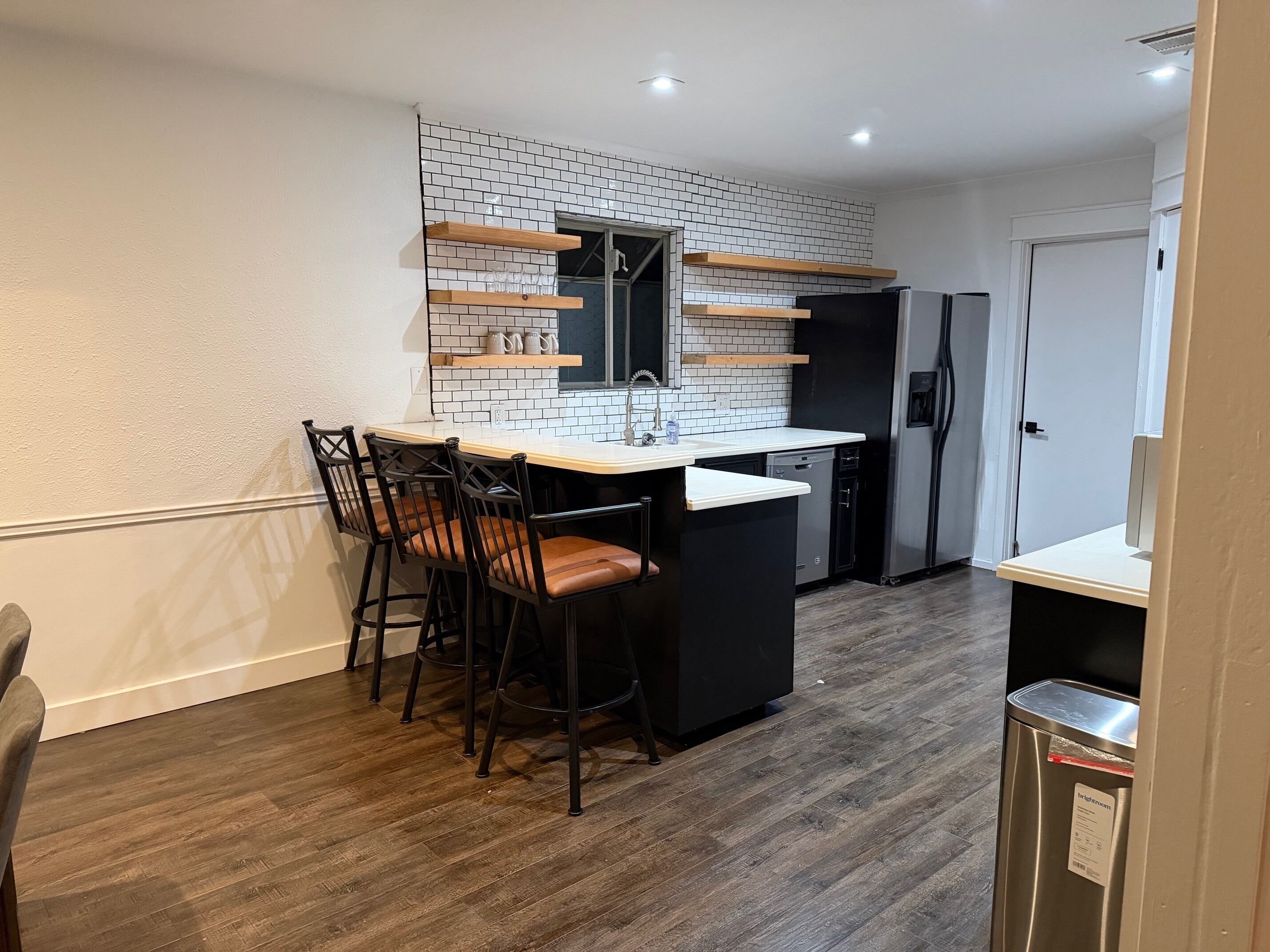 Une cuisine compacte avec un crédence carrelée de couleur blanche, un réfrigérateur noir, un lave-vaisselle et un comptoir-bar en bois avec des tabourets.