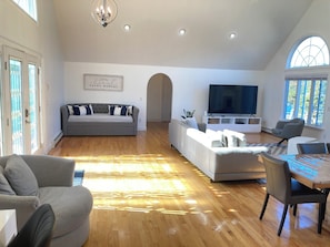 A spacious living room with a grey sofa, a dining table with chairs, a flat-screen TV, and a large window. - Elegant + relaxing + water views + Cape Cod experience (Bourne)
