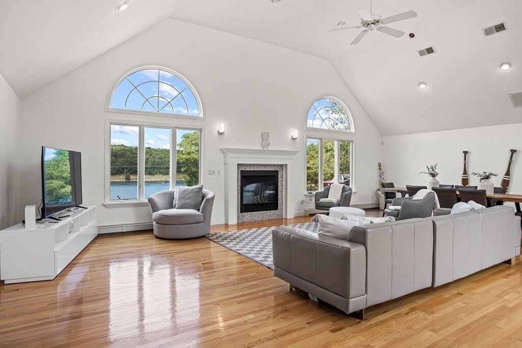 A spacious living room with a fireplace, large windows, and a ceiling fan.