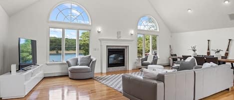A spacious living room with a fireplace, large windows, and a ceiling fan.