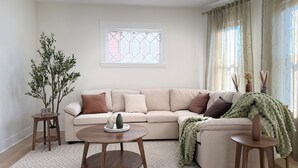 A living room with a beige sectional sofa, a round wooden coffee table, and a large window with sheer curtains. - Stylish 3BR Revere Home|Sleeps 8|Near Boston&Beach (Revere)