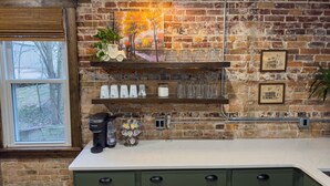 A kitchen with a brick wall, a window with blinds, a coffee maker, a kettle, and a shelf with cups and a plant. - Twin Bricks Railhouse near Route 66 (Bourbon)
