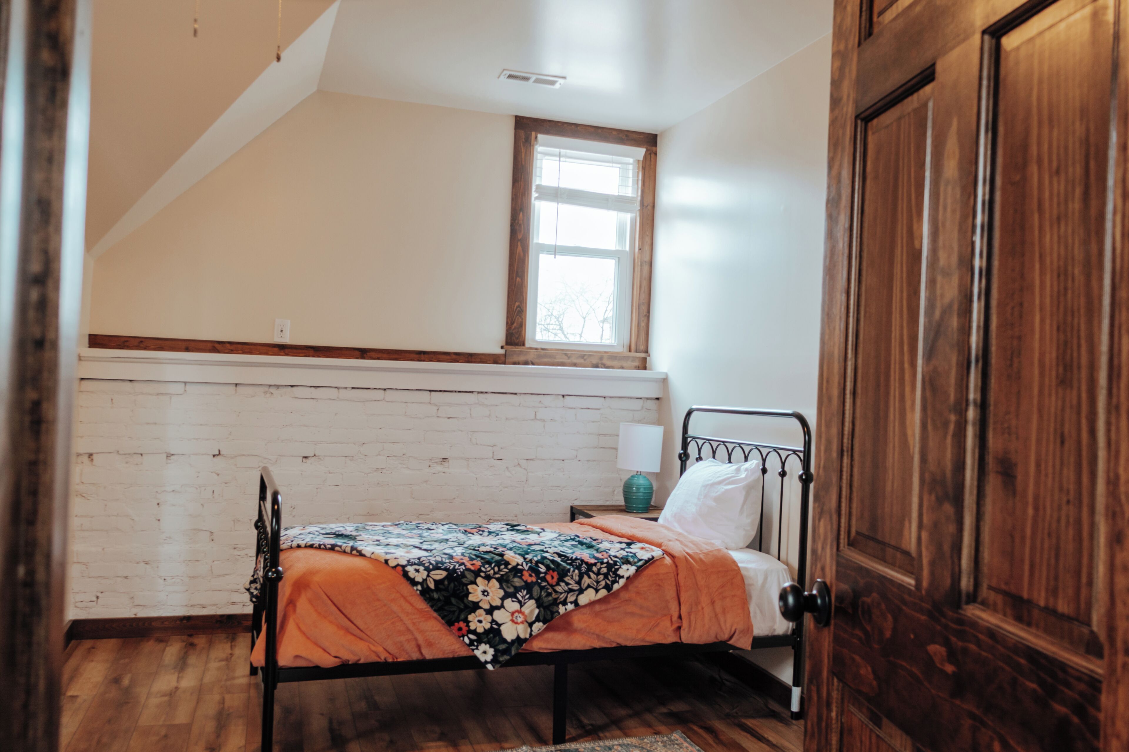 A bedroom with a sloped ceiling, a window, a bed with a floral bedspread, a nightstand with a lamp, and a wooden wardrobe.