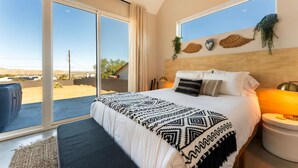 A bedroom with a large bed, a nightstand, a lamp, and a view of the desert through large sliding doors. - NEW: Sol + Shadow | Pool | Spa | Walk to Downtown (Joshua Tree)