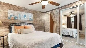 Une chambre à coucher avec un lit, un ventilateur de plafond, un tableau au mur et une porte-fenêtre coulissante donnant sur une autre pièce.