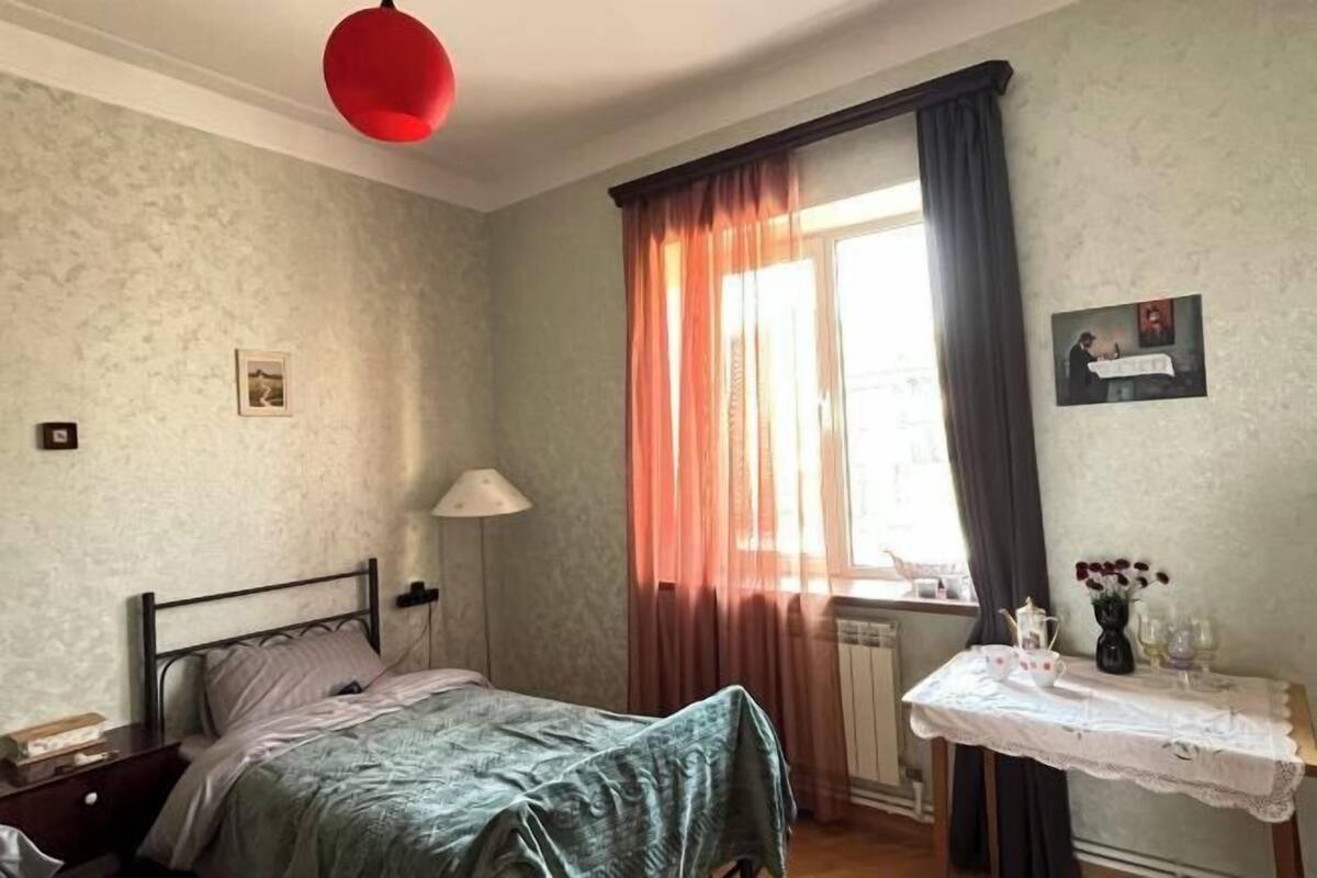 A bedroom with a bed, a nightstand, a table with a vase, and a window with curtains.