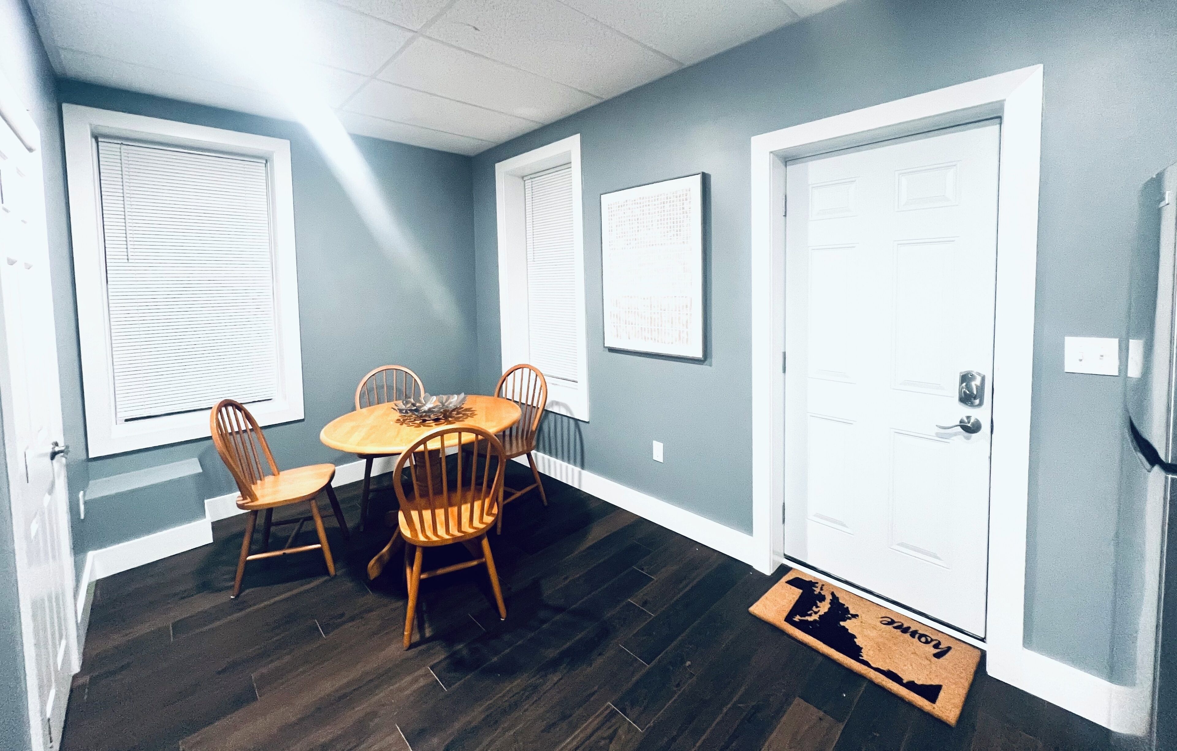 A dining area with a round table and four chairs, a white door, and a window with blinds.
