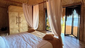 Quarto com cama, piso de madeira, uma janela grande e vista para a praia.