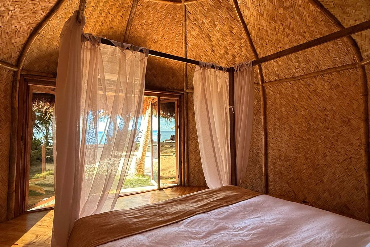 A room with a bed, a view of the beach, and a thatched roof.