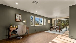 Una habitación amplia con suelo de madera, un escritorio con silla, una mesa de comedor con sillas y ventanas grandes que dan a vistas de árboles.