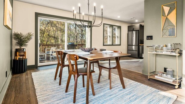 Un comedor moderno con una mesa grande, sillas y una araña. Se ve el exterior a través de amplias ventanas.