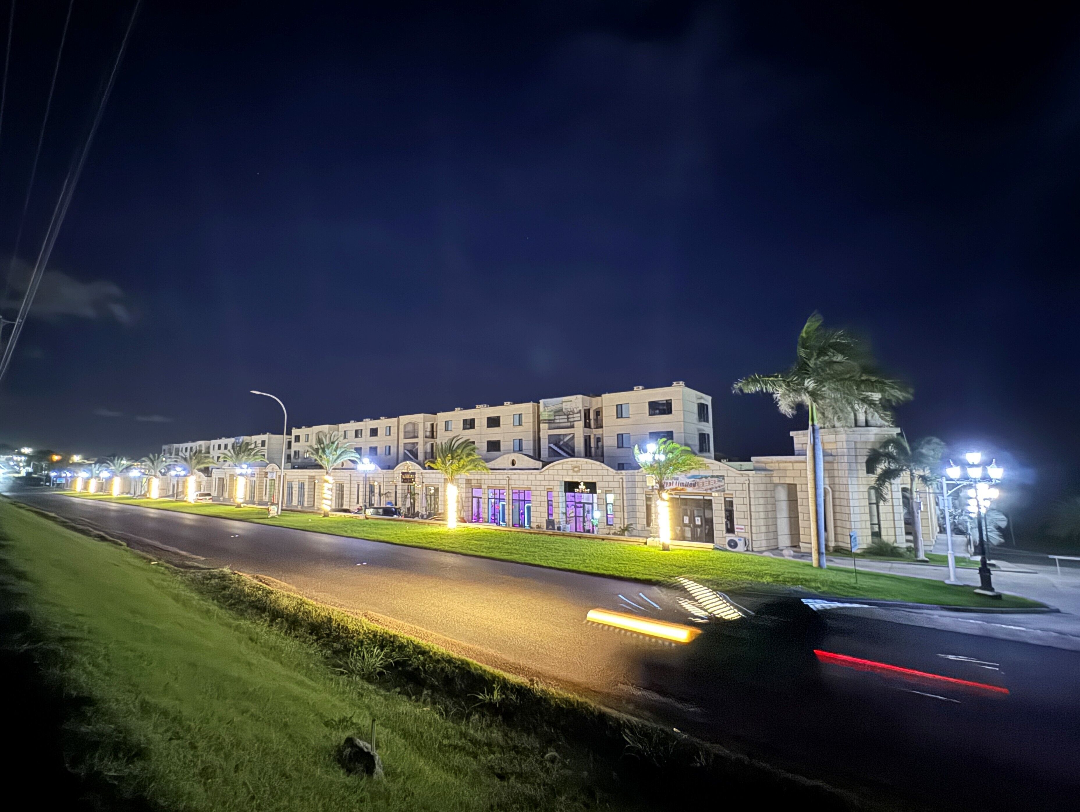 Un hotel de varios pisos con una fachada bien iluminada, un jardín ajardinado en la parte delantera y una calle con vehículos en movimiento.