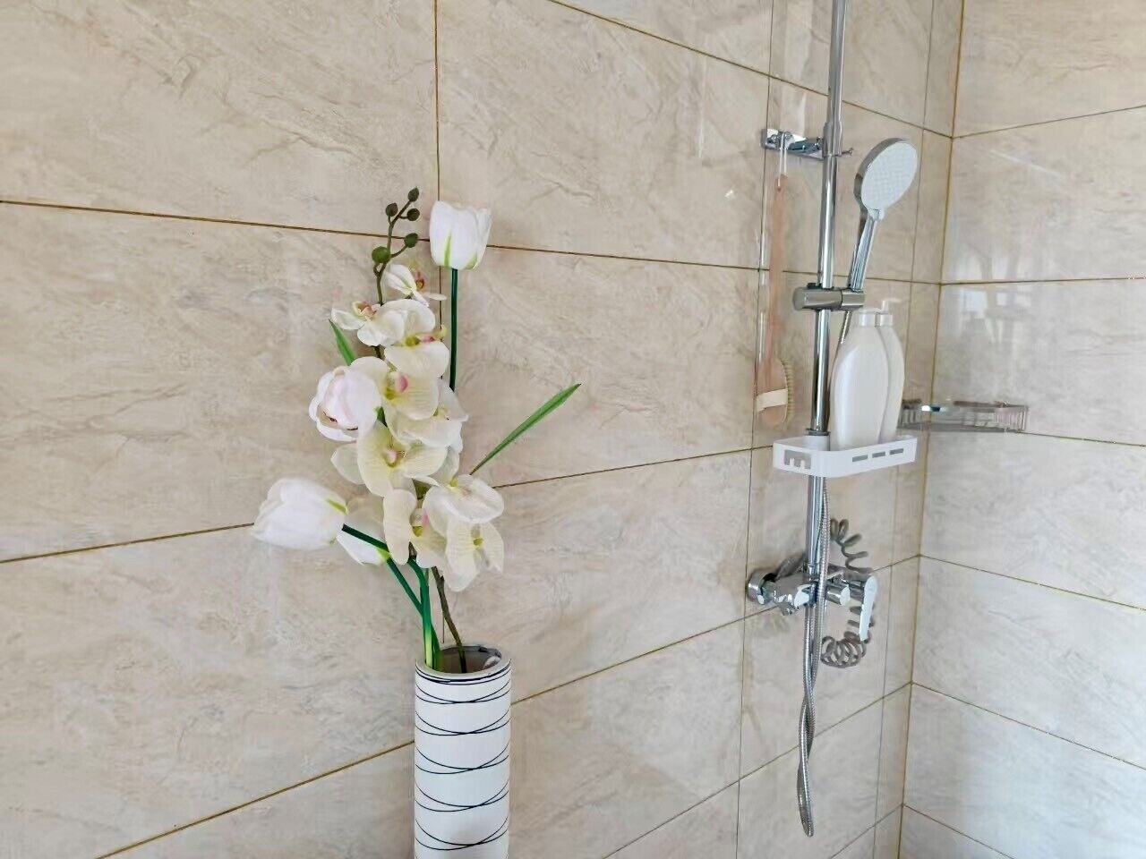 Une salle de bain avec un mur en marbre, une douche équipée d’un pommeau de douche à main et un vase rempli de fleurs blanches.