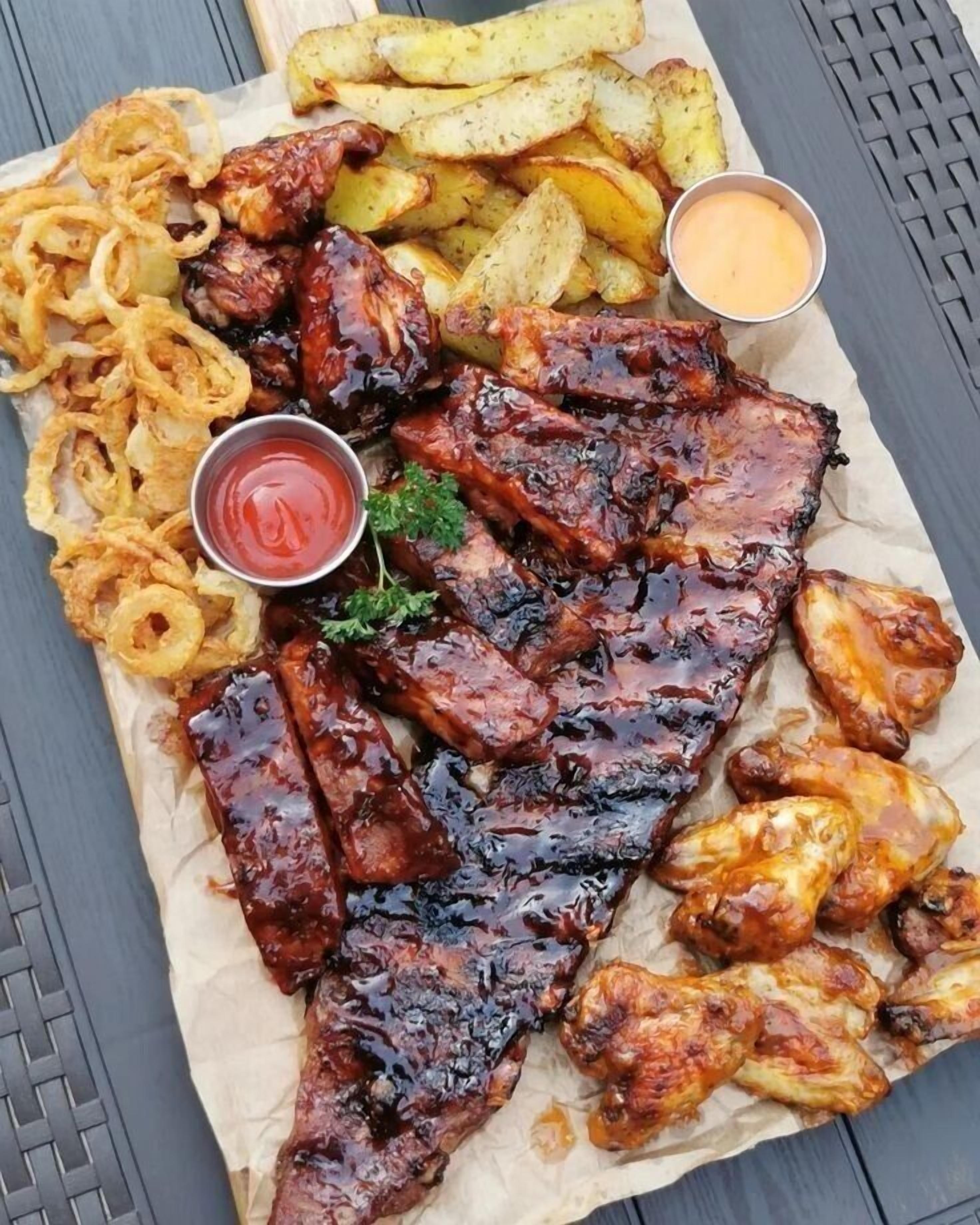 A meal consisting of ribs, wings, fries, onion rings, and two dipping sauces on a table.