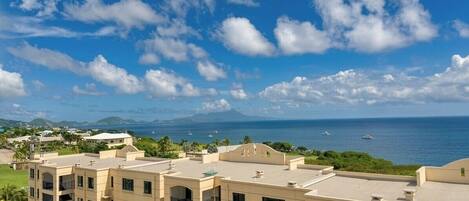 A multi-story hotel with balconies, a view of the ocean, and distant mountains.