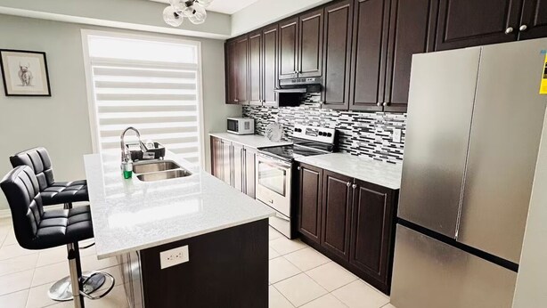 A modern kitchen with dark cabinets, a stainless steel refrigerator, a built-in oven, and a large island with a sink. - Comfy home sleeps 13+ guest with wifi &parking Netflix (Brampton)