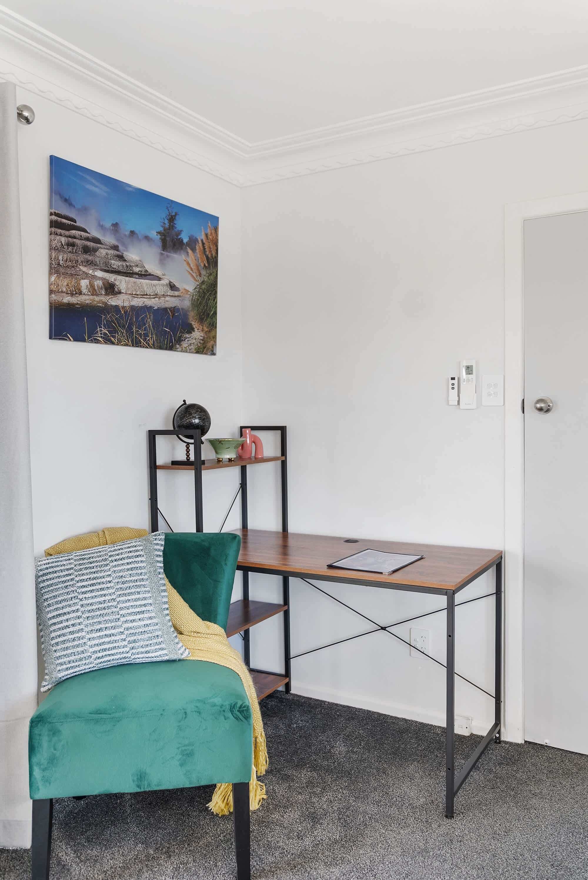 A corner room with a green chair, a small wooden table, a wall-mounted shelf with decorative items, and a painting of a natural landscape.