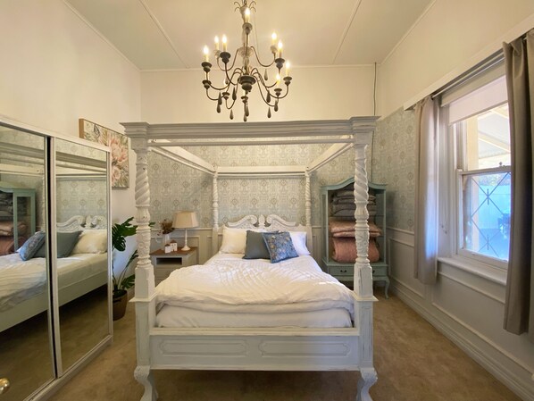 Un lit à baldaquin surmonté d’un lustre, un grand miroir et une fenêtre avec des rideaux.