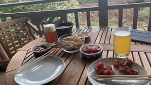 Une table en bois sur laquelle se trouvent une assiette de sashimi, un verre de bière et des bols de légumes marinés, le tout sur un balcon offrant une vue sur la verdure et un paysage rocheux.