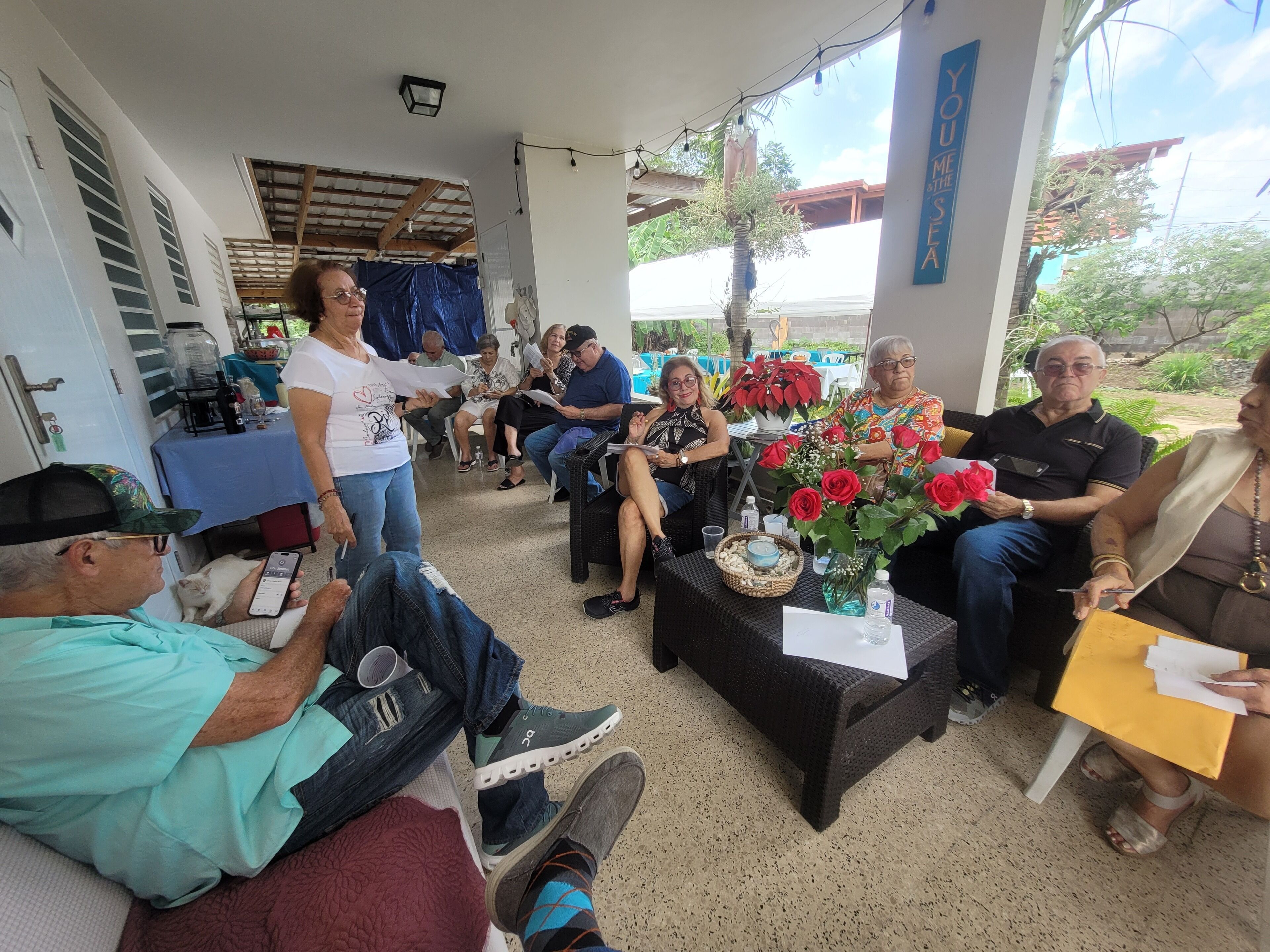 A group of people seated on outdoor furniture in a covered area with a table and chairs, a small table with a vase of flowers, and a banner on the wall.