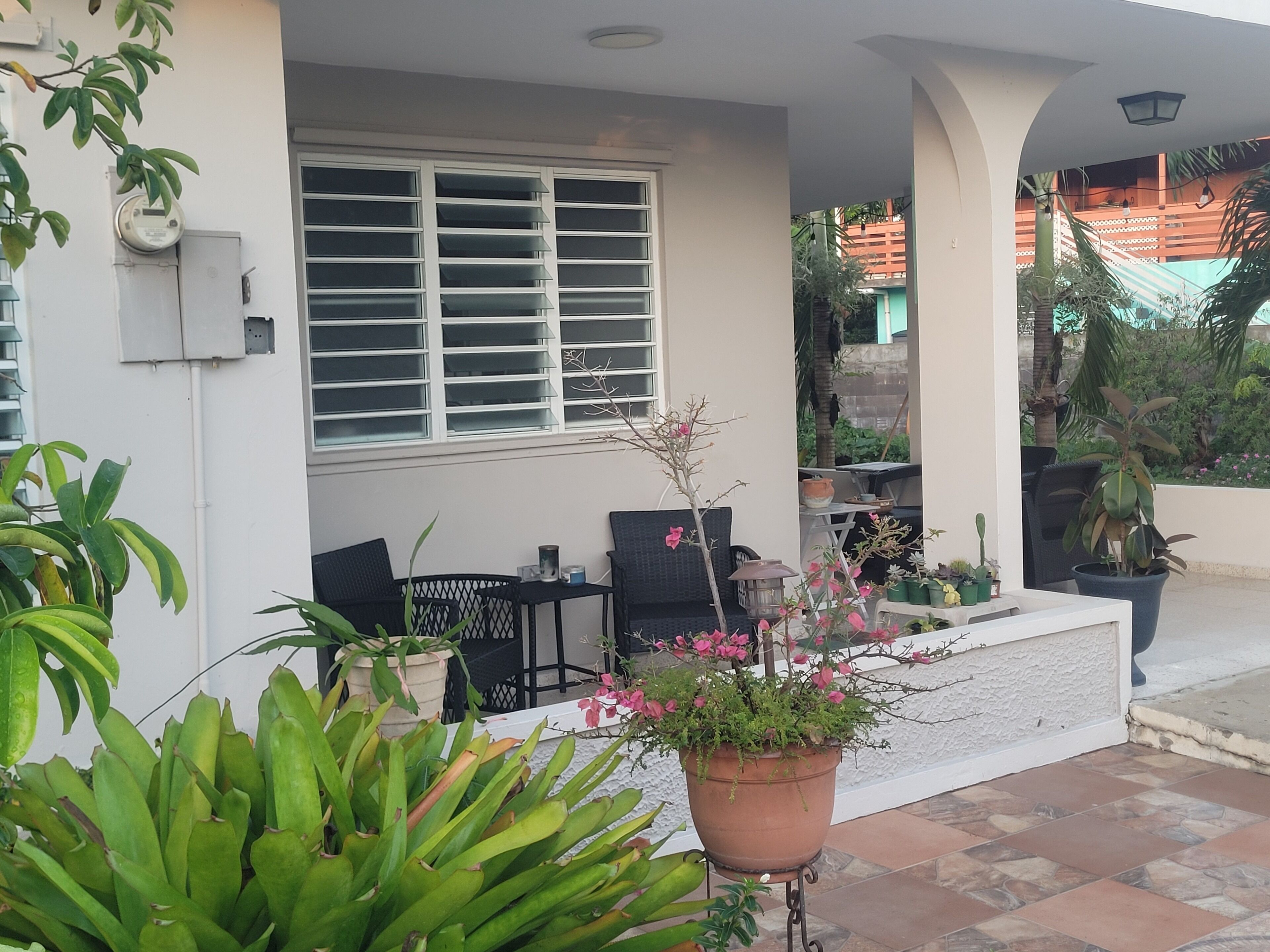 A covered outdoor seating area with a table and chairs, surrounded by potted plants and a window with blinds.