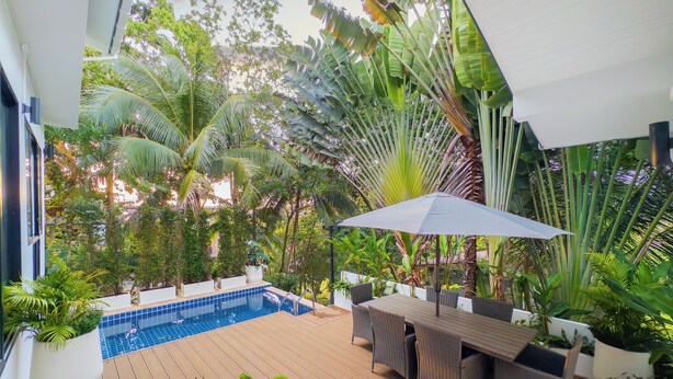 Zona junto a la piscina con terraza de madera, mobiliario exterior y exuberante vegetación tropical.
