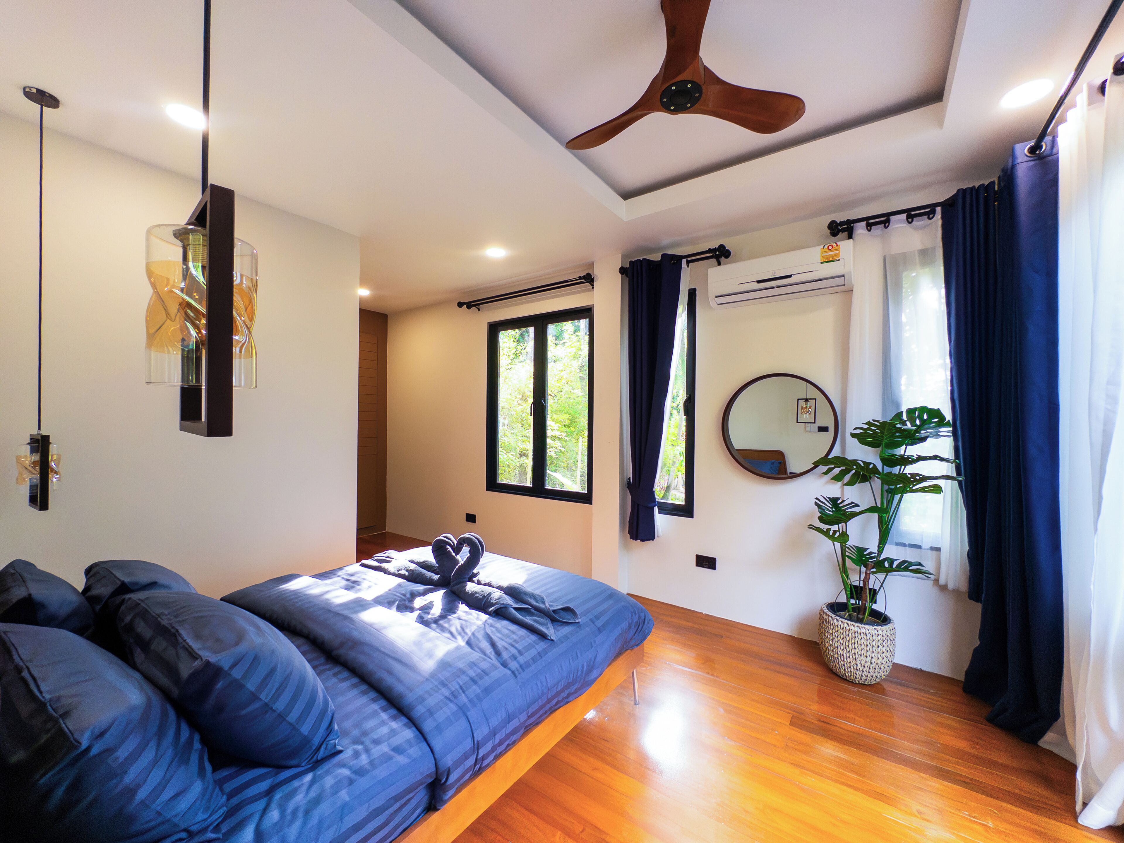 Une chambre à coucher avec un lit, un plancher en bois, un ventilateur de plafond, une fenêtre avec des rideaux, une plante en pot et un miroir fixé au mur.