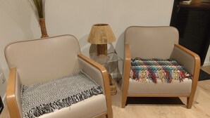 Dos sillones con cojines de diferentes estampados, una mesita con una lámpara y un jarrón alto con follaje verde.