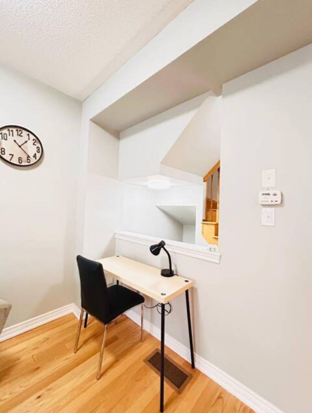 A small, well-lit room with a wooden desk, a black chair, a wall clock, and a white ceiling.