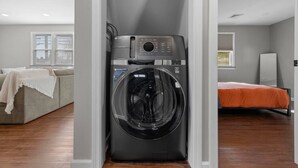 A compact laundry room with a front-loading washer and dryer, a gray sofa with a white throw blanket, and a bed with an orange bedspread. - #22- 4 Br 2 Ba house-Central CT (New Britain)
