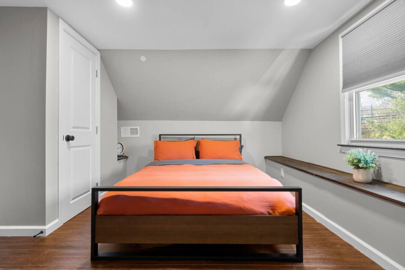 Une chambre à coucher avec un lit, une fenêtre avec des stores, une porte et une petite étagère sur laquelle se trouve une plante.