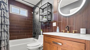 A modern bathroom with a toilet, bathtub, wooden vanity, and a mirror with a light. - #22- 4 Br 2 Ba house-Central CT (New Britain)