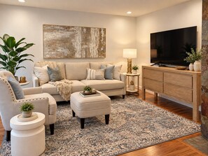 A modern living room with a sofa, armchair, ottoman, and a flat-screen TV.