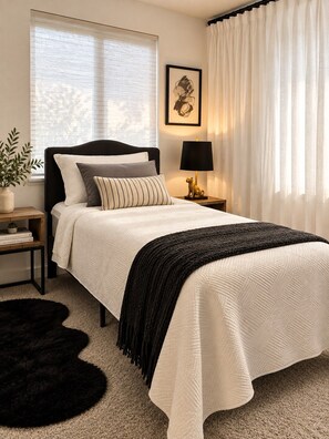 A neatly made bed with a white duvet, a black throw blanket, and several pillows. There is a small bedside table with a lamp and a decorative item. A large window with sheer curtains allows natural light into the room.