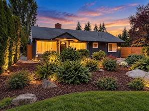 A residential house with a dark exterior, a well-maintained garden with various plants and rocks, and a clear sky. - The Guest House - Newly Remodeled (Lynnwood)