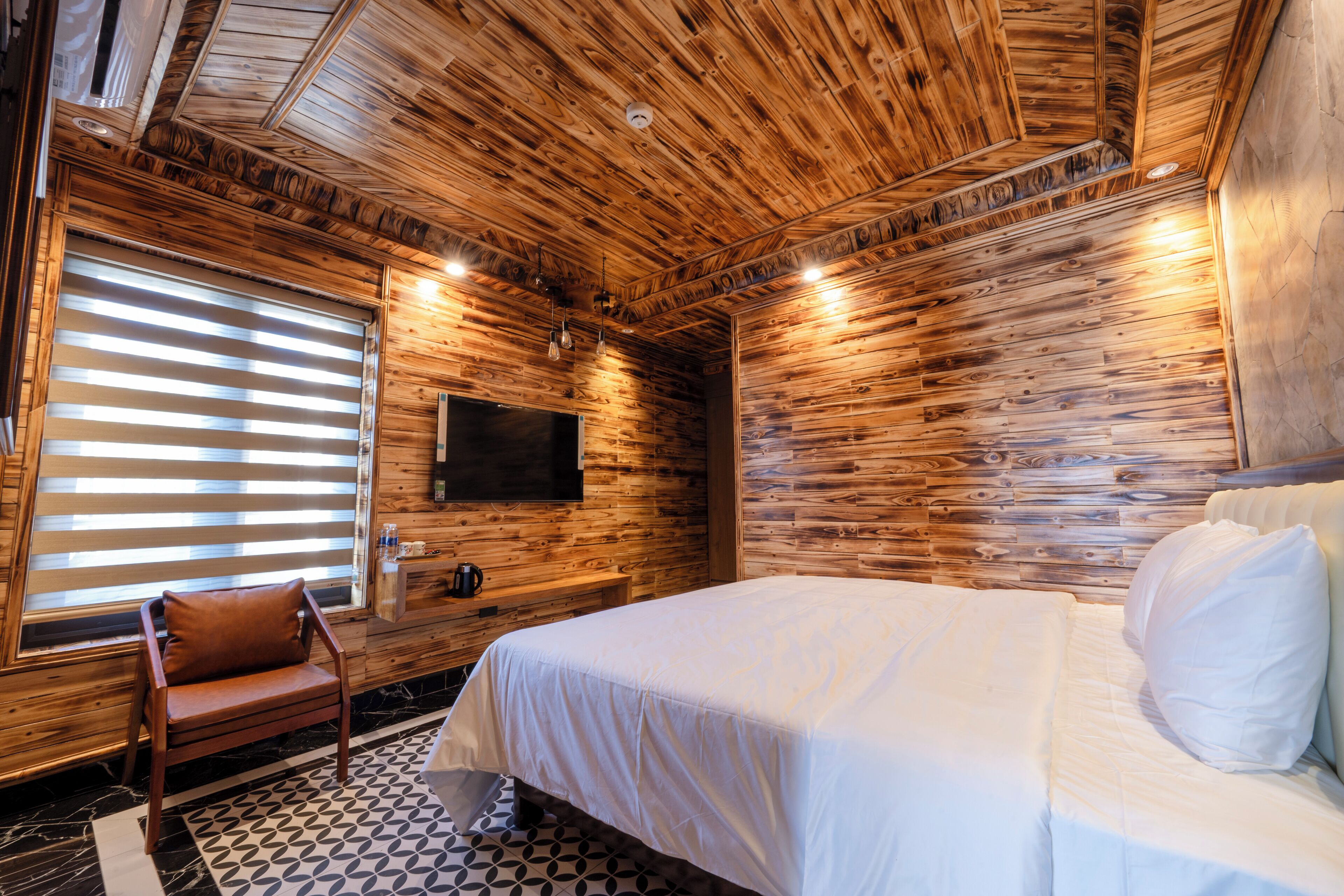 A wooden-paneled room with a bed, a chair, a TV, and a window with blinds.