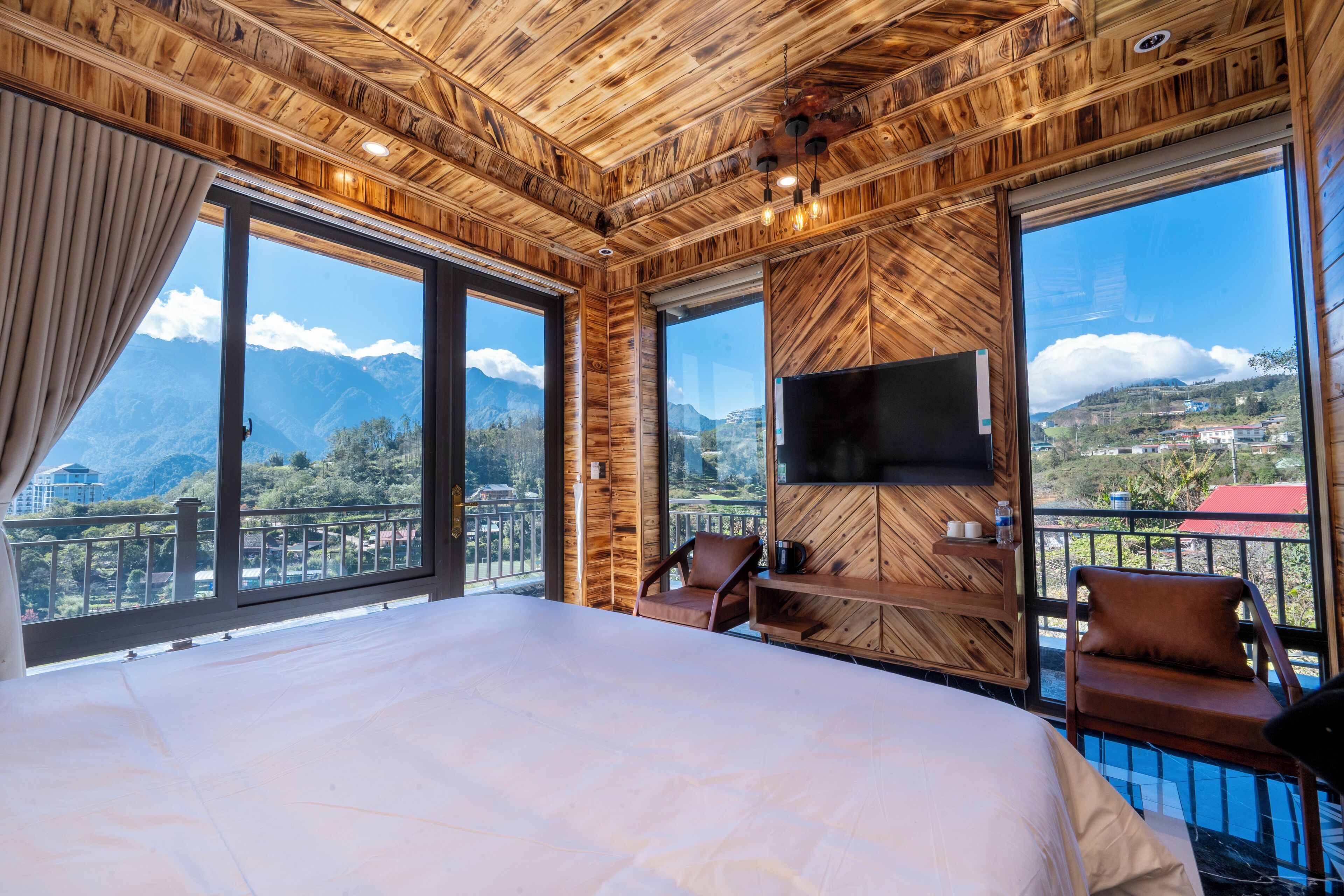 A large bed in a room with wooden walls and a TV mounted on the wall. There are two chairs and a window with a view of mountains.
