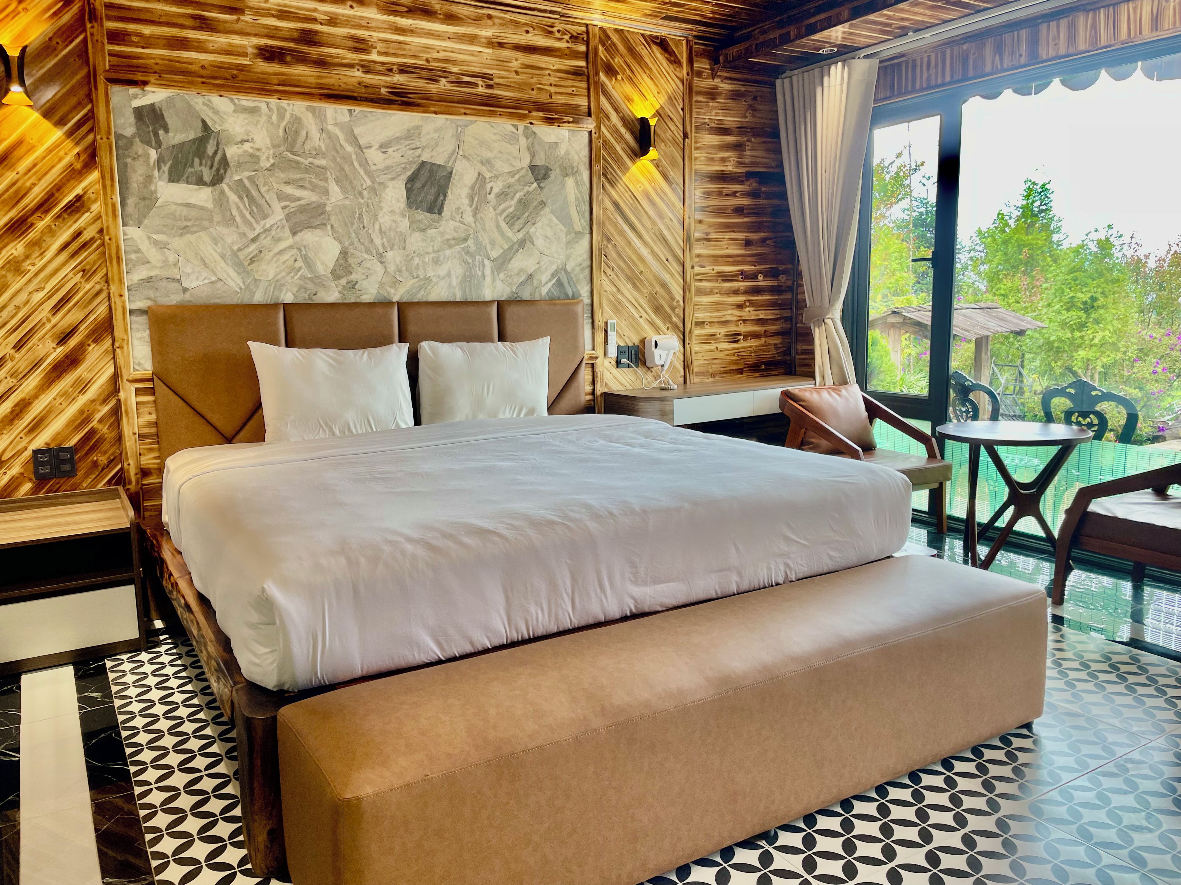 A hotel room with a large bed, a wooden headboard, a stone-patterned wall, and a view of greenery outside.