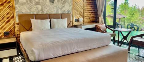 A hotel room with a large bed, a wooden headboard, a stone-patterned wall, and a view of greenery outside.