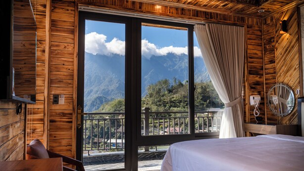 Habitación de cabaña de madera con cama, una silla, un ventanal con cortinas y un balcón con vistas a montañas.