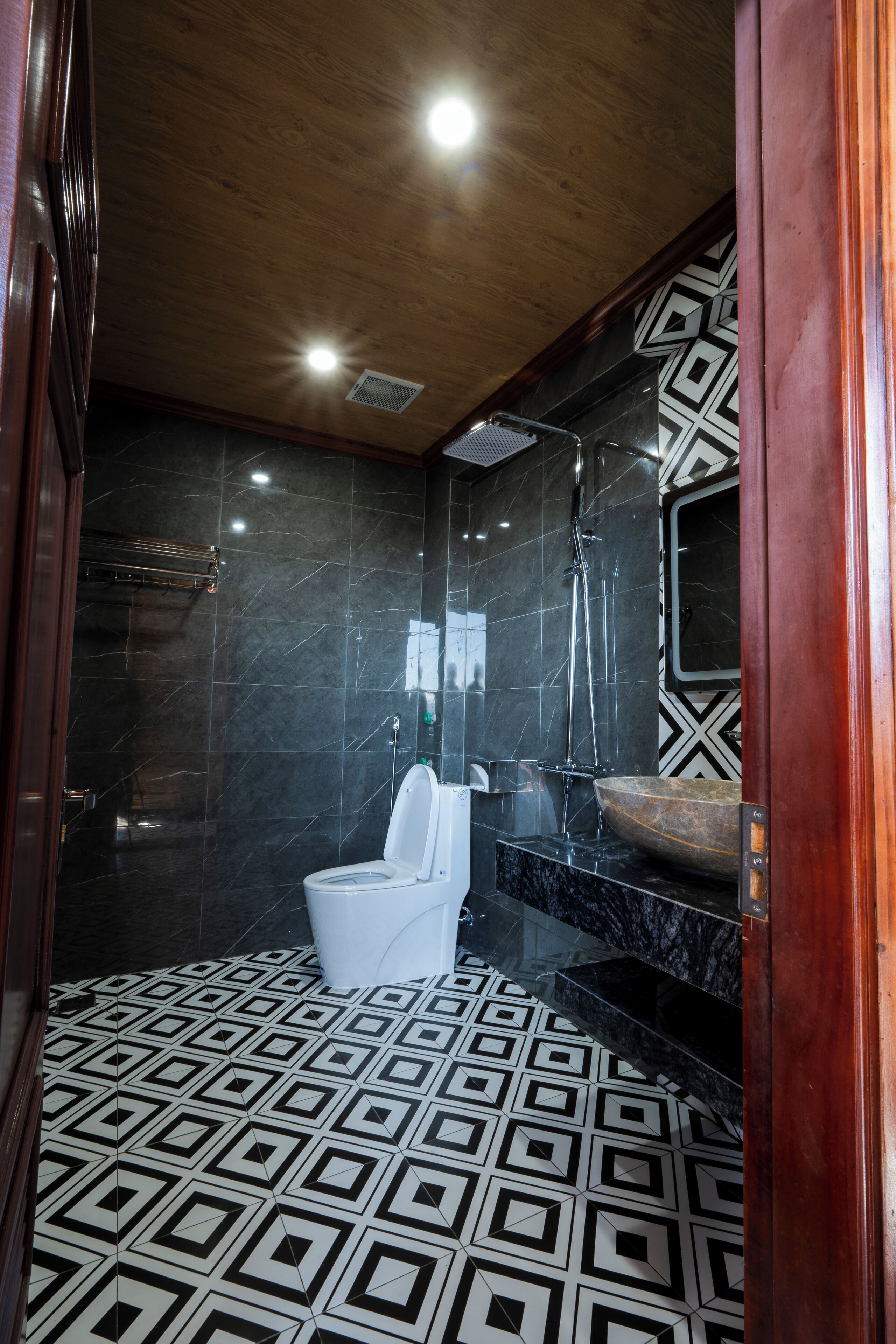 A modern bathroom with a geometric patterned floor, a white toilet, a black marble sink, and a wooden ceiling.