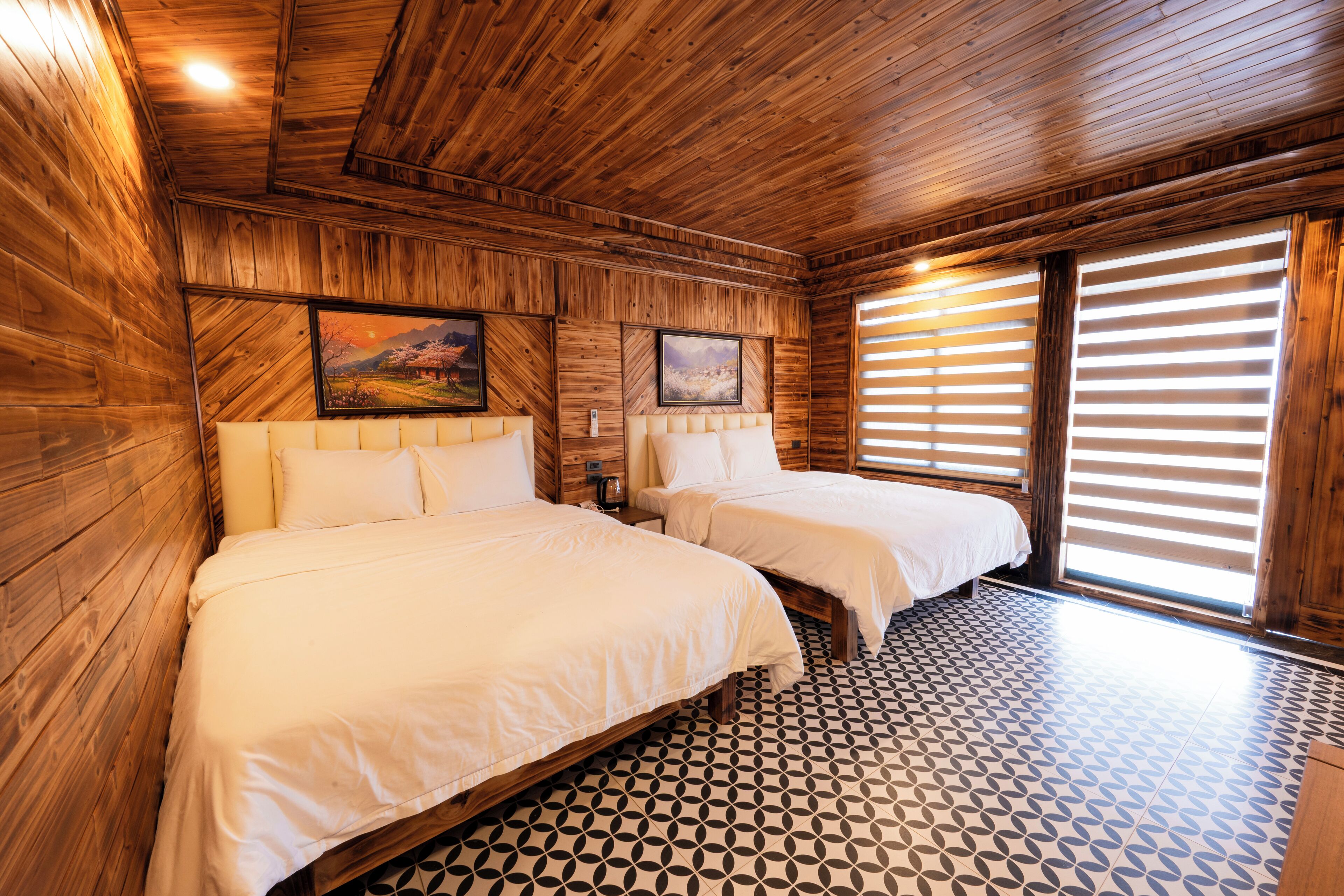 A wooden-paneled room with two beds, a patterned floor, and a large window with blinds.