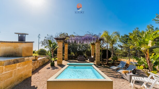 Una piscina con una pérgola y sillas de descanso, rodeada de follaje y un muro de piedra.